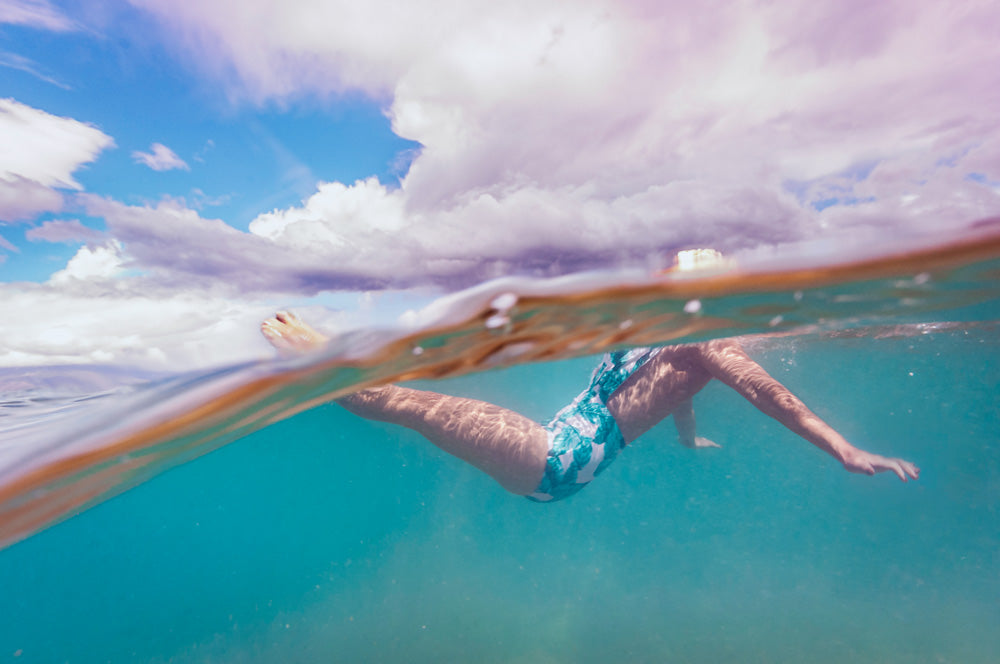 Female swimmer in retro style floral bathing suit is immersed in ocean, split-view with sky and clouds in background.